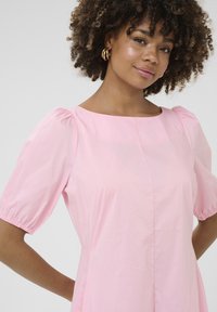 Robe s puff sleeves iz svetlo roza tkanine, ki ima okroglo ovratnico in šiv po sprednji strani. Gladka tekstura s nabranimi akcenti na rokavih.