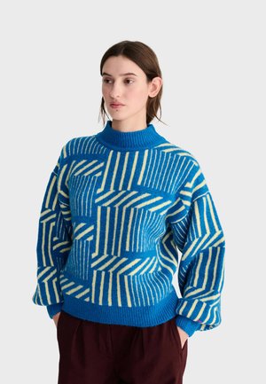 Jeune femme portant un pull à motif géométrique bleu et blanc avec des manches larges et un pantalon bordeaux, debout devant un fond uni.