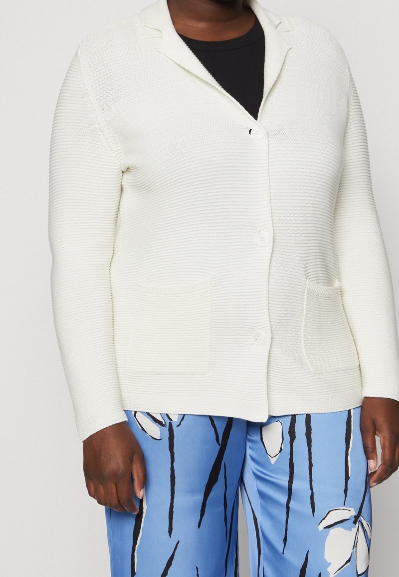 Persona con un cárdigan blanco texturizado y abotonado sobre una blusa negra, combinado con pantalones azules que presentan un patrón floral abstracto en blanco y negro.