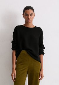 Schwarzer Strickpullover mit umgeschlagenen Bündchen, getragen über olivgrünen Cordhosen. Einfaches Design, runder Ausschnitt und strukturierte Stoffdetail.