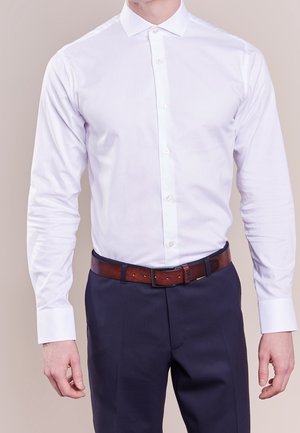 Homme portant une chemise blanche cintrée boutonnée et rentrée dans un pantalon bleu marine foncé avec une ceinture en cuir marron, debout devant un fond uni.