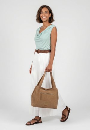 Mujer sonriente con camiseta sin mangas de color verde claro, falda blanca plisada, cinturón marrón y sandalias, sosteniendo un gran bolso marrón claro.