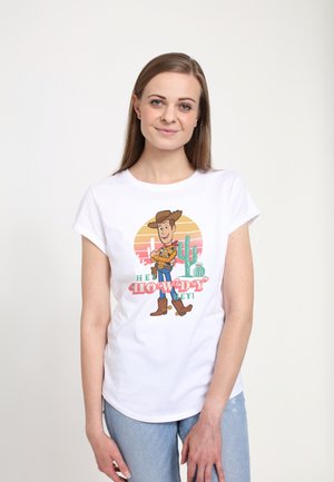 Vrouw draagt een wit T-shirt met Woody uit Toy Story, een cactus en de tekst "Hey Howdy Hey!", staand tegen een effen lichte achtergrond.