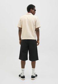 Chemise à manches courtes rayée en crème et rose clair, associée à un short noir. Portée avec des baskets noires et des chaussettes blanches. Fond uni.