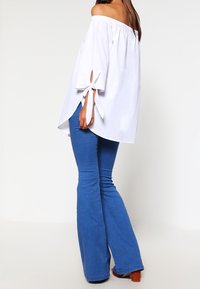Blusa bianca scollata sulle spalle con maniche lunghe, caratterizzata da dettagli con fiocco e vestibilità comoda, abbinata a jeans blu a zampa.