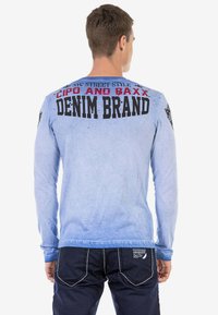 Langärmliges blaues Hemd mit dunkelblauen Akzenten, das auf der Rückseite bedruckten Text aufweist. Der Stoff wirkt weich mit einer abgestuften Textur und lässiger Passform.