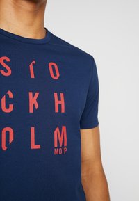 Marineblå bomuld T-shirt med lodret rød tekst, der staver "STOCKHOLM" og "MO’P" i en fed, moderne skrifttype. Korte ærmer og rund halsudskæring.