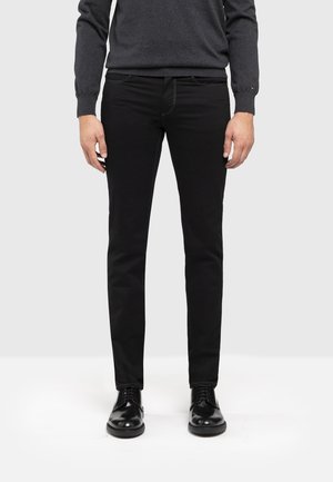Mann trägt schwarze Slim-Fit-Hose, schwarze Lederschuhe und einen dunkelgrauen Langarmpullover, steht vor einem schlichten Hintergrund.