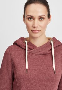 Haut à capuche bordeaux avec une finition texturée, conception de cou croisé et cordons crème. Présente des accents en métal au niveau de la capuche.