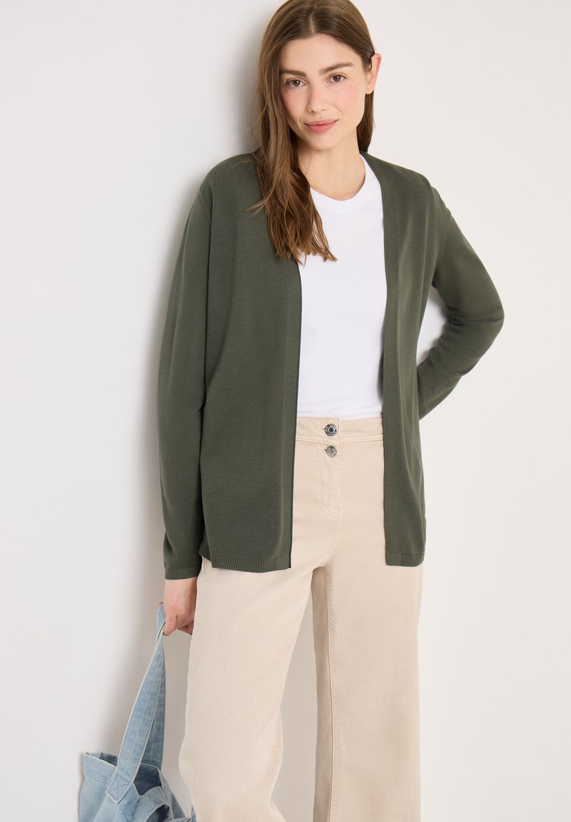 Jeune femme portant un cardigan ouvert vert olive, un t-shirt blanc, un pantalon beige, tenant un sac fourre-tout bleu clair, debout devant un fond uni.