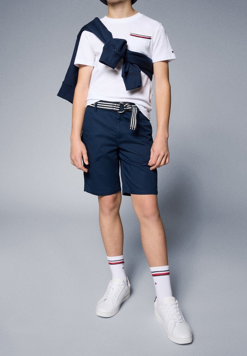 Personne portant un t-shirt blanc, un short bleu marine avec une ceinture rayée, des baskets blanches, des chaussettes blanches avec des rayures rouges et noires, et un sweat-shirt bleu marine posé sur l'épaule.