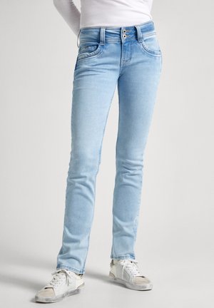 Personne portant un jean slim bleu clair et des baskets blanches, debout devant un fond uni gris clair.