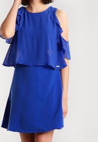 Vestido azul con superposición de volantes en capas y diseño de hombros fríos. Hecho de una tela liviana, presenta un escote redondo clásico y falda acampanada.