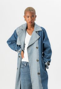 Denim trench coat featuring a two-tone design, large collar, double-breasted buttons, and front pockets. Soft denim texture in blue shades.