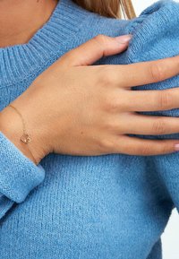 Goldene Kettenarmband mit einem herzförmigen Anhänger, getragen an einer Hand. Die Trägerin trägt einen blauen Strickpullover und hat hellfarbene Nägel.