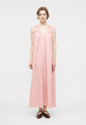 Nő egyenesen állva, ujjatlan, hosszú rózsaszín ruhában és sötétbarna Mary Jane cipőben, egyszínű világos háttér előtt.