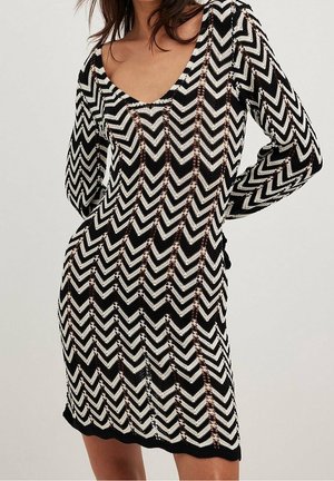 Femme portant une robe en maille noire et blanche à motif zigzag, manches longues, encolure dégagée, posant les mains sur les hanches devant un fond uni.