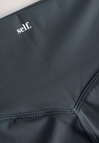 Črne atletske legice iz gladkega blaga, z izbočeno šivom in tiskanim logotipom "self." v belem na območju pasu.