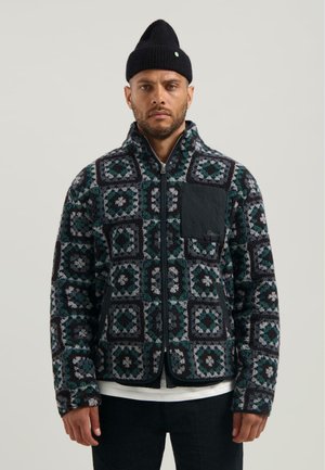 Hombre con chaqueta de forro polar estampada con cremallera frontal, bolsillo en el pecho negro, gorro negro, camisa blanca y pantalones negros sobre un fondo liso.