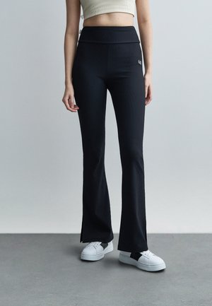 Γυναίκα που φοράει μαύρο παντελόνι καμπάνα με ριμπ ύφανση και λογότυπο στο πλάι, καθώς και λευκά αθλητικά παπούτσια, στέκεται σε γκρι δάπεδο μπροστά σε λευκό φόντο.