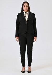 Schwarzer maßgeschneiderter Anzug mit einem taillierten Blazer, der über einen einzelnen Knopf und angeschnittene Revers verfügt, kombiniert mit einer schmal geschnittenen Hose und einem weißen Hemd mit Kragen.