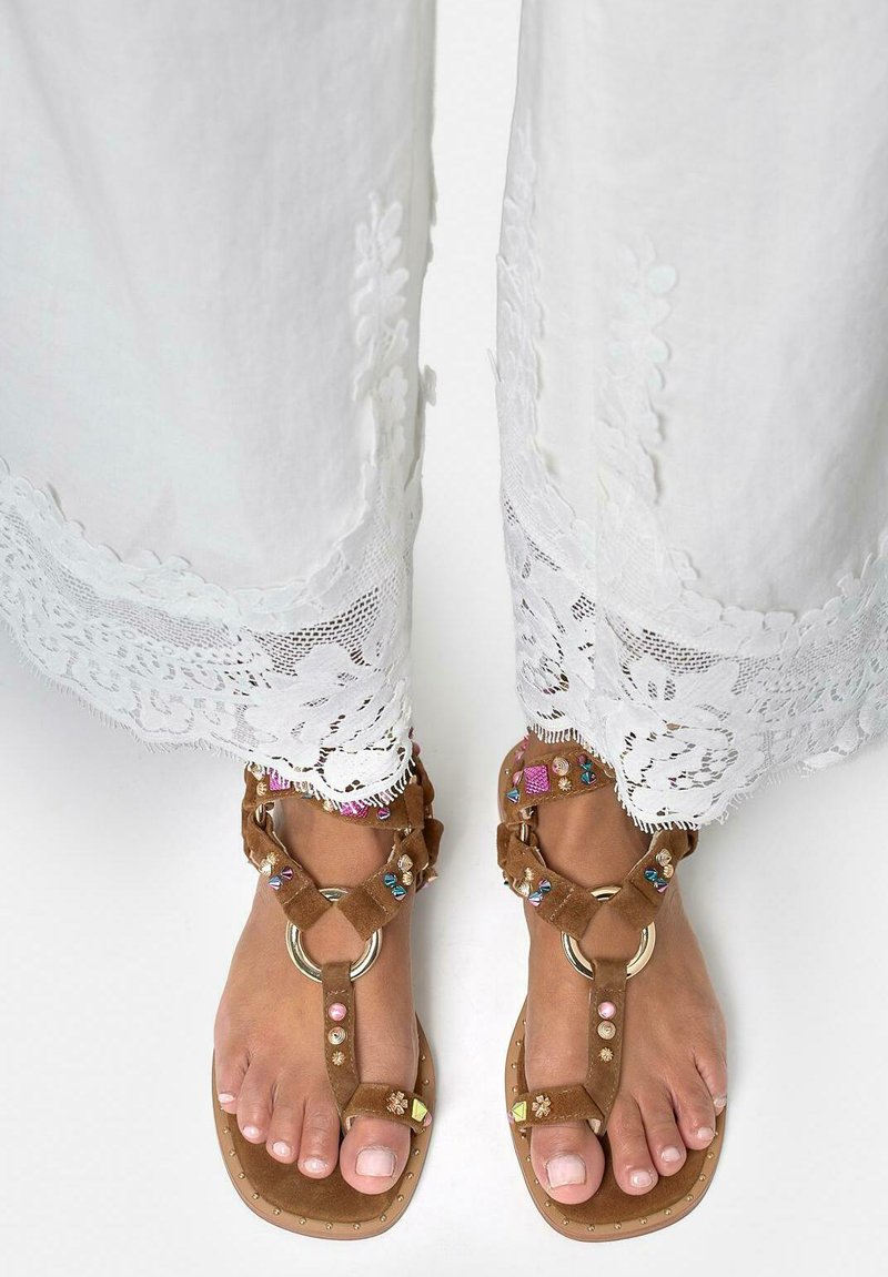 Stopy w brązowych zamszowych sandałach z kolorowymi koralikami i złotymi pierścionkami, w zestawieniu z białymi spodniami z koronkowymi lamówkami.