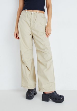 Persona con pantalones beige de pierna ancha con cintura elástica y zapatos negros tipo slip-on con plataforma y perforaciones, de pie sobre un fondo blanco.