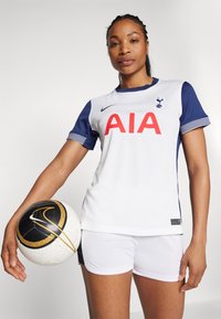 Witte voetbalkleding met marineblauwe mouwen, rood "AIA" logo en teamembleem. Gecombineerd met witte shorts en houdt een zwart-wit voetbalsbal vast.