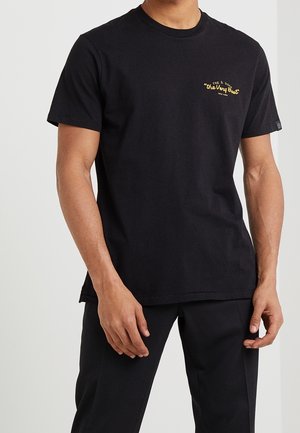 Mann trägt ein schwarzes kurzärmeliges T-Shirt mit gelbem Text "the Very Best" auf der linken Brustseite und schwarze Hose.