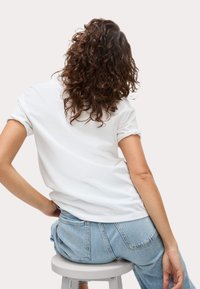 T-shirt bianca a maniche corte con risvolti sulle maniche, indossata con jeans a vita alta di colore azzurro chiaro; modella seduta con capelli ricci vista da dietro.