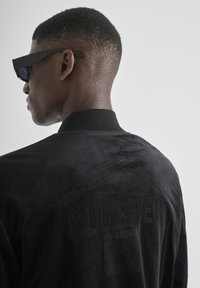 Chaqueta bomber negra con tela texturizada, con la palabra "CLUBBER" en relieve en la parte trasera, combinada con gafas de sol negras y angulares.