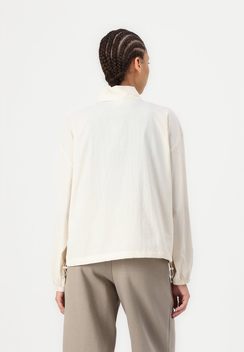 Veste crème claire avec un col, coupe ample et manches à poignets élastiques. Présente des accents à cordon de serrage à l'ourlet et un tissu texturé.