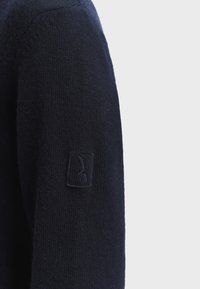 Tummansininen villaneule, jossa on teksturoitu neulekuvio ja hienovarainen logopainatus vasemmassa hihassa. Läheltä otettu kuva hihan yksityiskohdasta.