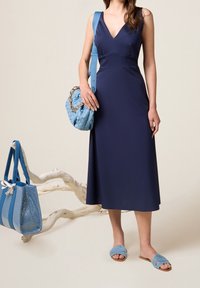 Robe sans manches de couleur marine avec un col en V, accompagnée d'un sac à main tissé bleu et de sandales à glisser bleues. Se distingue par son tissu lisse et ses lignes épurées.