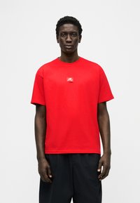 Άνδρας που στέκεται προς τα εμπρός, φορώντας φωτεινό κόκκινο κοντομάνικο μπλουζάκι με μικρό λευκό λογότυπο στο στήθος και μαύρο παντελόνι.