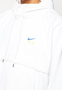 Valkoinen huppari, jossa on sininen Nike-logo ja keltainen "AIR"-teksti. Valmistettu pehmeästä kankaasta, jossa on säädettävät nyörit.