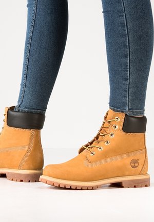 Tan leather ankle boots with black padded collars worn with blue skinny jeans, shown on a white background.