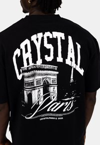 Czarny bawełniany t-shirt z dużym białym napisem "CRYSTAL" oraz grafiką Łuku Triumfalnego, z napisem "Paryż" napisanym poniżej.
