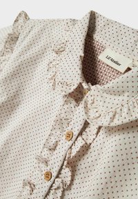 Blusa leggera in tessuto con base crema e piccoli motivi a diamante rossi, colletto volant e bottoni in legno lungo la parte anteriore.
