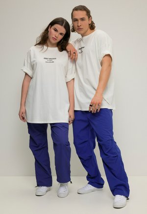 Oversized witte t-shirts met zwarte print, gecombineerd met blauwe cargobroeken. Het model draagt witte sneakers, met eenvoudige ontwerpen en minimale detailing.