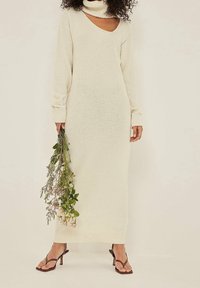 Abito lungo in maglia di colore avorio con colletto alto e dettagli a taglio. Maniche e orlo a coste. Il modello tiene un mazzo di fiori.