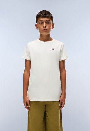 Un jeune homme porte un T-shirt blanc orné d’un petit logo du drapeau norvégien et un pantalon olive, sur fond bleu pâle.
