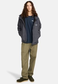 Herenjas met een marineblauw en grijs geruit patroon, ritssluiting en een capuchon. Draagt over een marineblauw shirt met olijfgroene cargobroek en donkere schoenen.