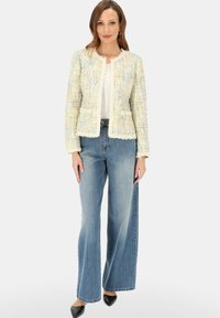 Kobieta ubrana w kremową i pastelową tweedową kurtkę o fakturze, biały top, szerokie niebieskie dżinsy i czarne buty z ostrym czubkiem, stojąca na jednolitym tle.