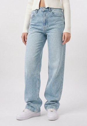 Kvinde iført lyseblå højtaljede vidde jeans, hvide sneakers og en cropped cremefarvet ribstrikket sweater, stående mod en ensfarvet baggrund.