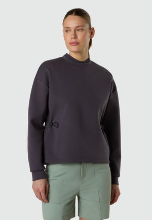 Femme portant un pull à manches longues foncé et un short vert clair, debout devant un fond uni, regardant sur le côté.