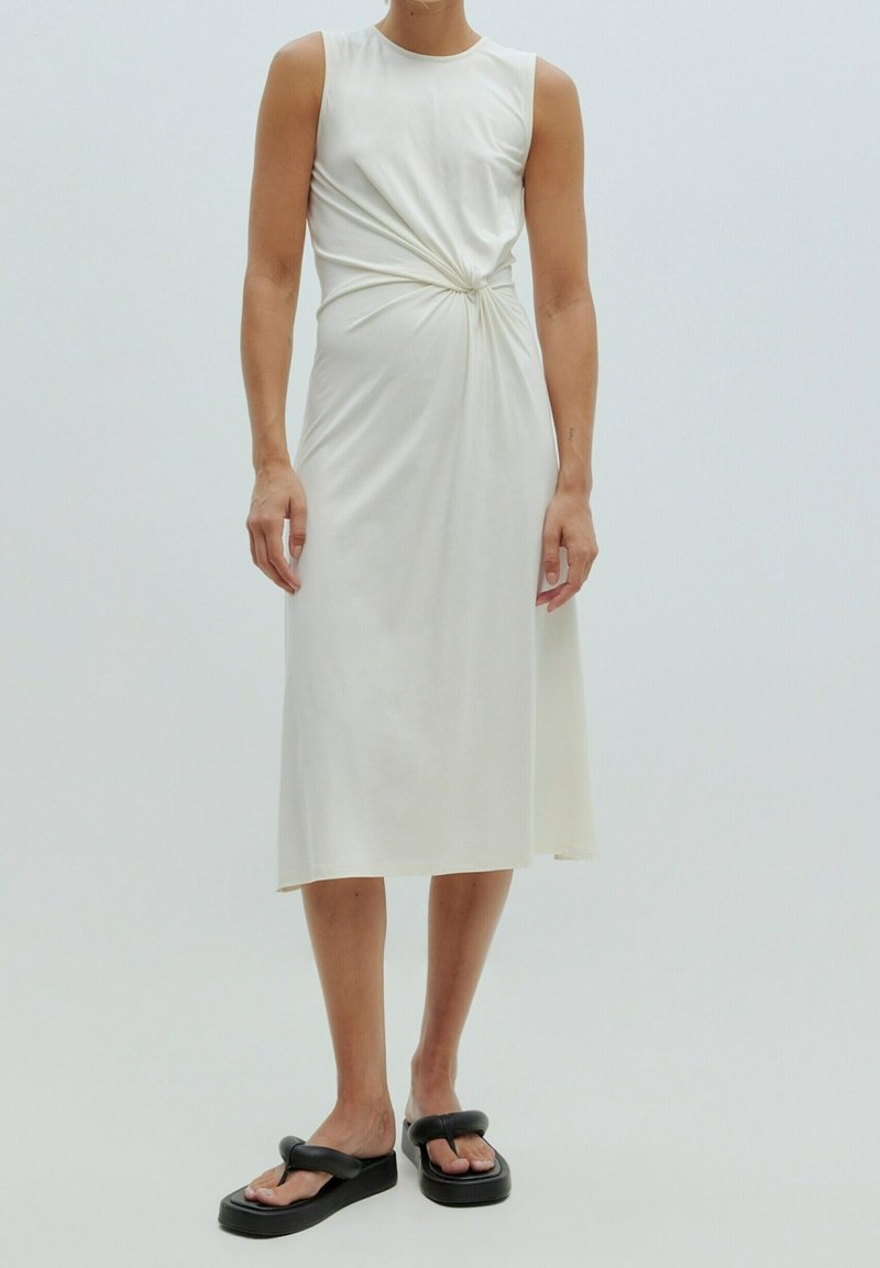 Robe midi blanche sans manches avec un détail de nœud à l'avant, portée avec des tongs à plateforme noires, sur une personne debout sur un fond uni.