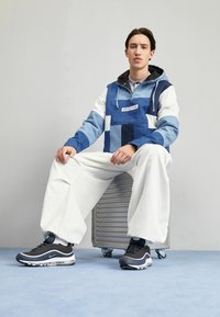 Sininen ja valkoinen tilkkutäkkihuppari tavanomaisella mitoituksella, valkoiset leveät housut ja mustat urheilukengät, joissa on ilmanvaimennus ja siniset yksityiskohdat.