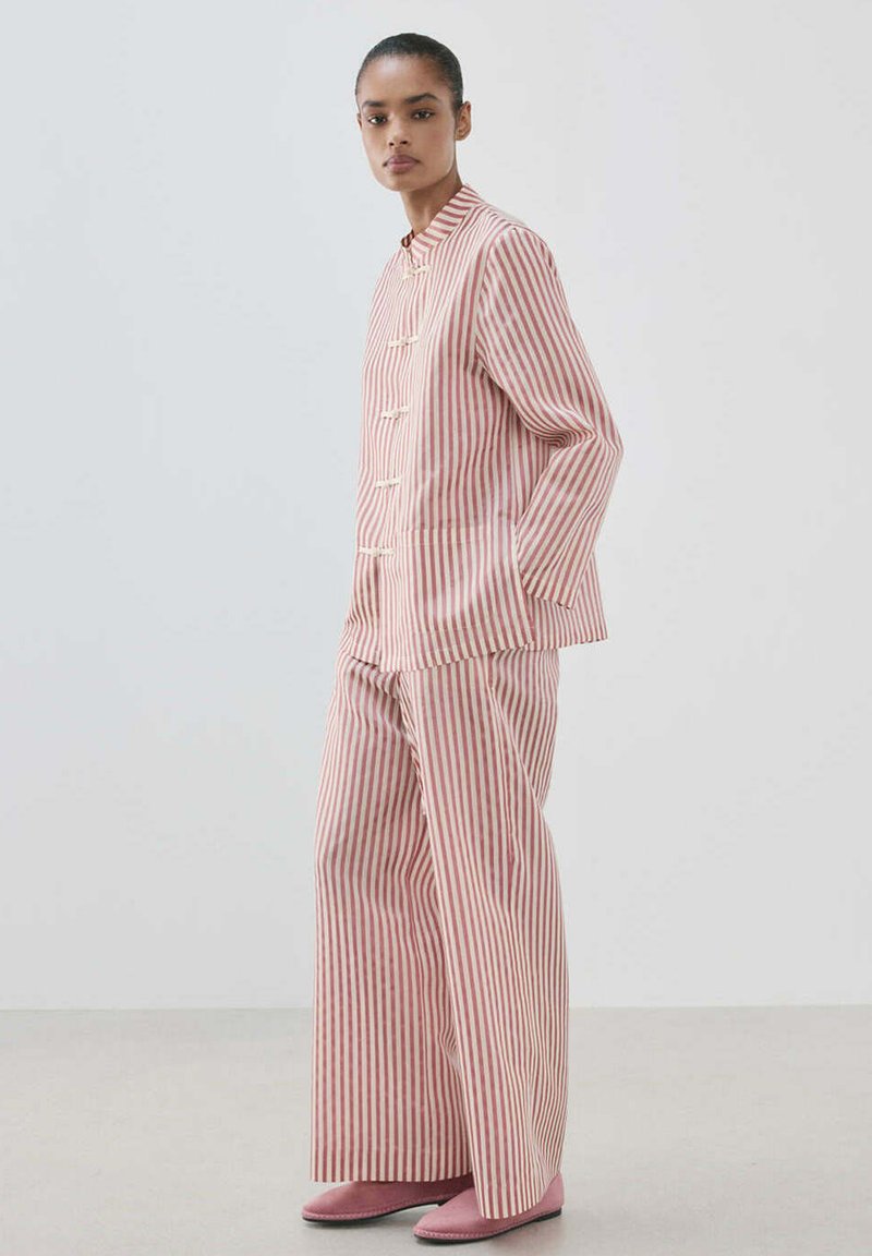 Femme portant un ensemble deux-pièces à rayures verticales rose et blanc avec une veste boutonnée et un pantalon à jambes larges, debout les mains dans les poches.
