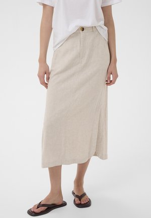 Personne portant une jupe midi beige avec une fente à l’avant, une chemise blanche manches courtes rentrée, et des tongs noires sur fond blanc.